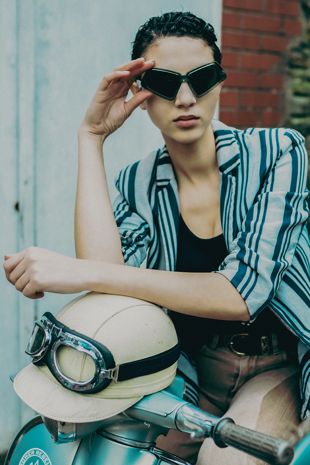 Model raising black Ffin MOD designer sunglasses beside a teal scooter, cream helmet and goggles