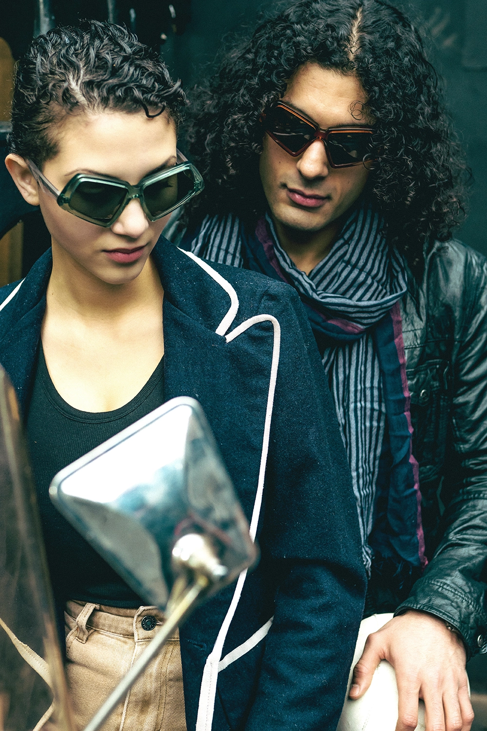Two models in dark angular Ffin MOD sunglasses beside a scooter mirror