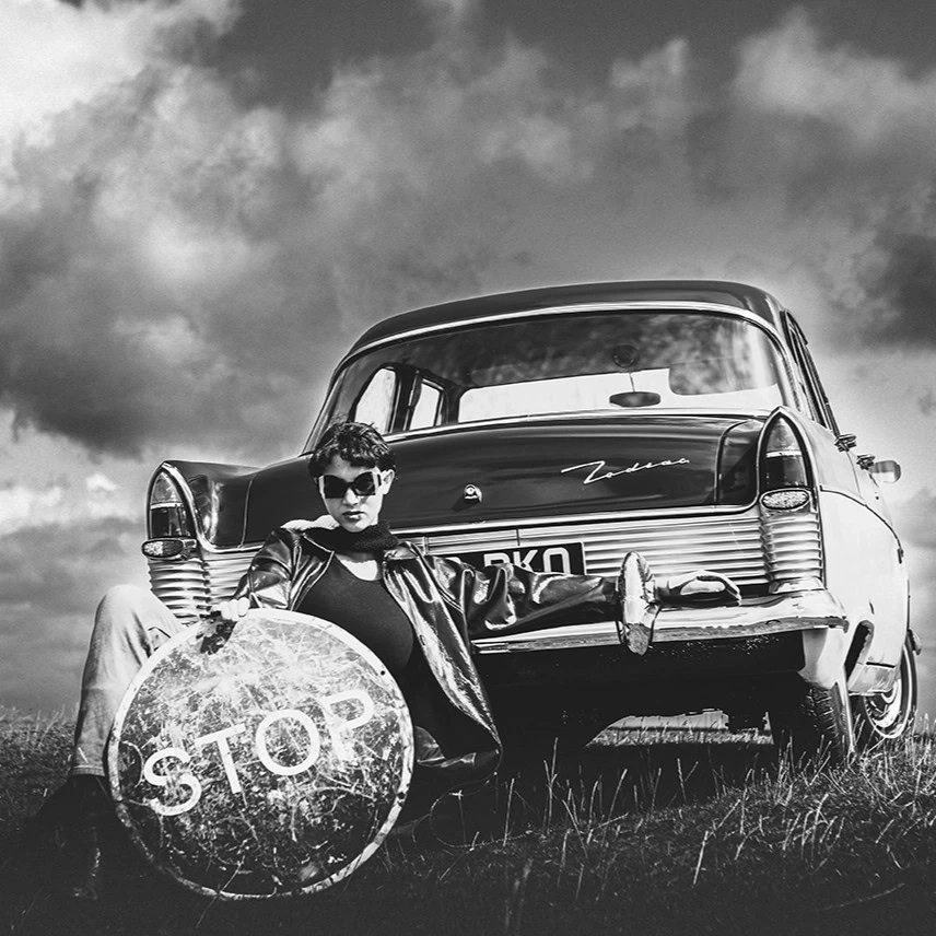 Model in Ffin sunglasses reclining against a vintage car on open moorland, holding a STOP sign
