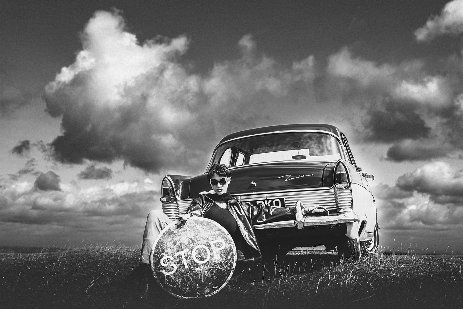 Model in sunglasses reclining against a vintage car on open moorland, holding a STOP sign, Ffin slow design shoot