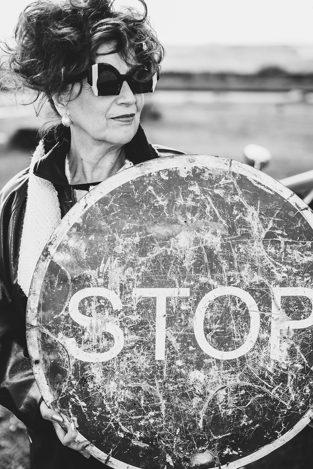 Older woman in angular black Ffin sunglasses holding a weathered STOP sign, slow design shoot