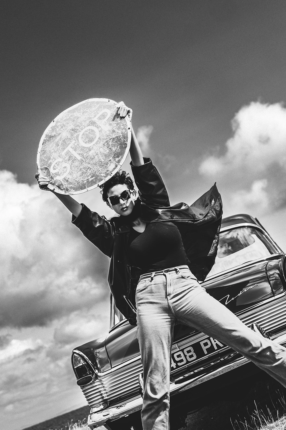 Model in Ffin sunglasses leaping beside a vintage car, holding a STOP sign overhead