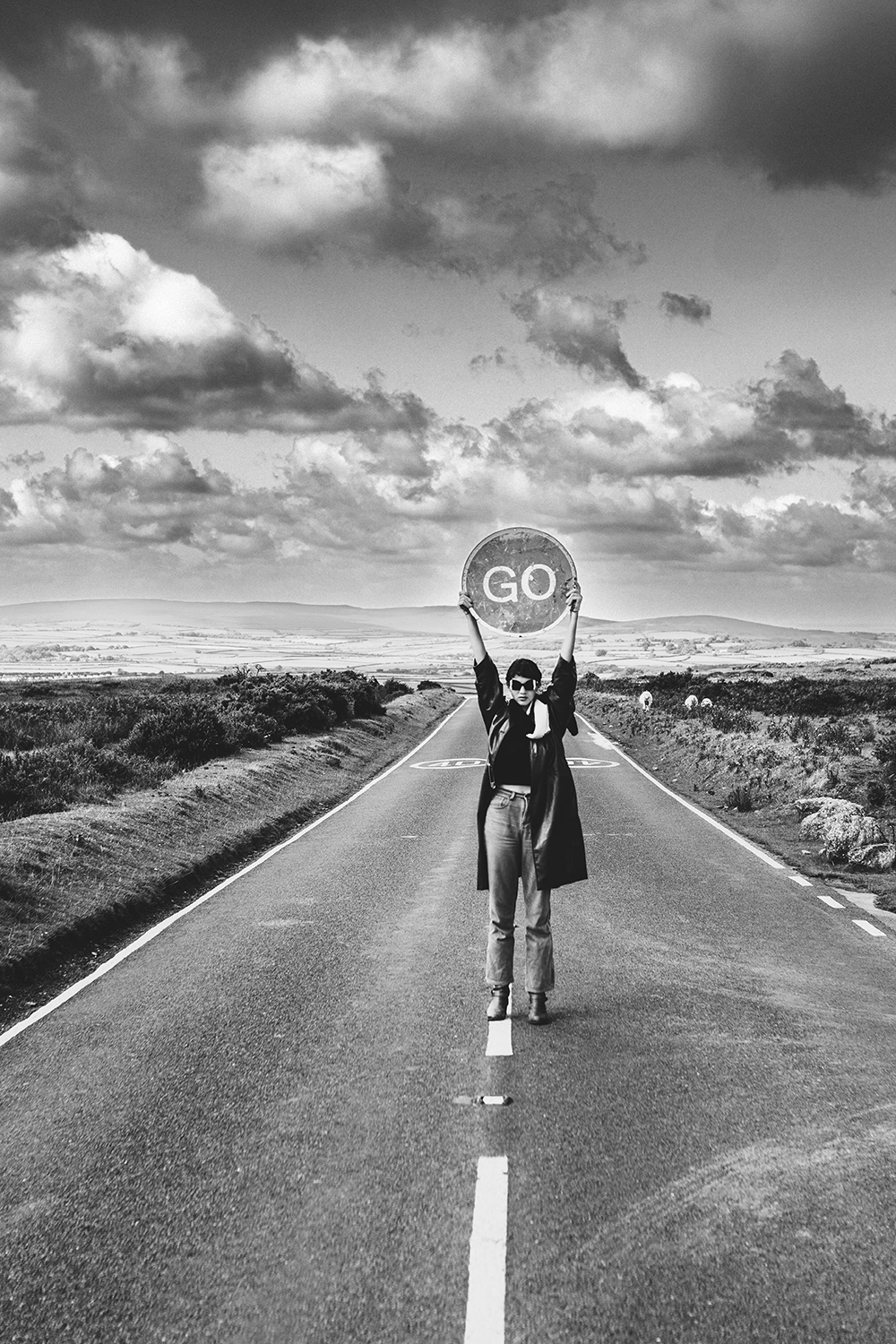 Figure holding a GO sign aloft on an empty moorland road, Ffin slow design shoot