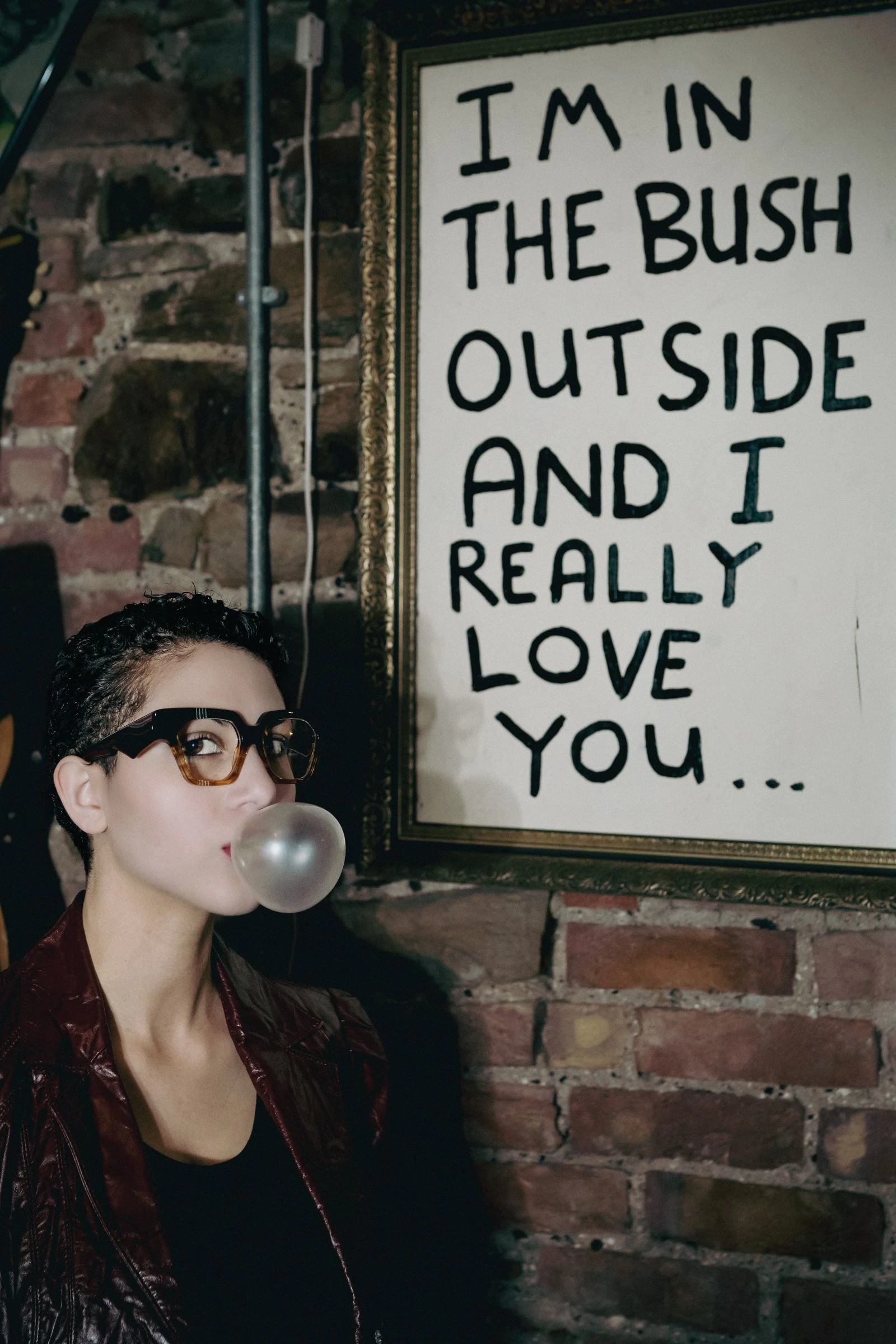Woman in black Ffin AMO optical frames blowing a bubble beside a sign reading "I'm in the bush outside and I really love you"