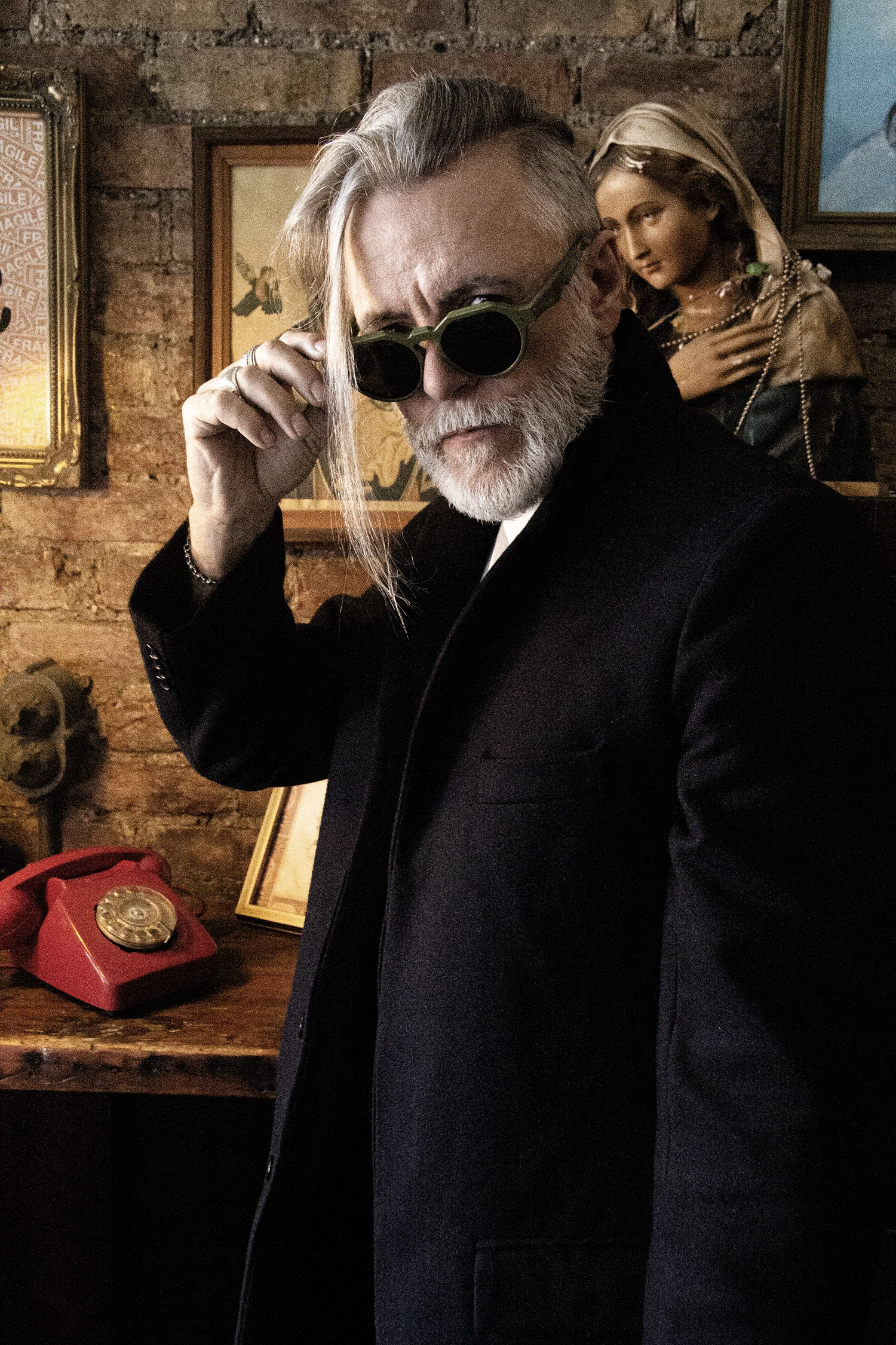 Steve Balsamo lowering olive Ffin NON designer sunglasses on his nose, a red telephone behind him