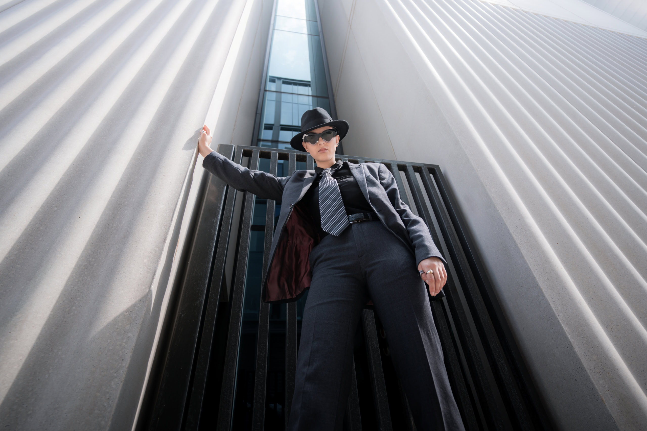 Riannah in a suit and fedora wearing Ffin NOS sunglasses, one hand on a fluted building, shot from below
