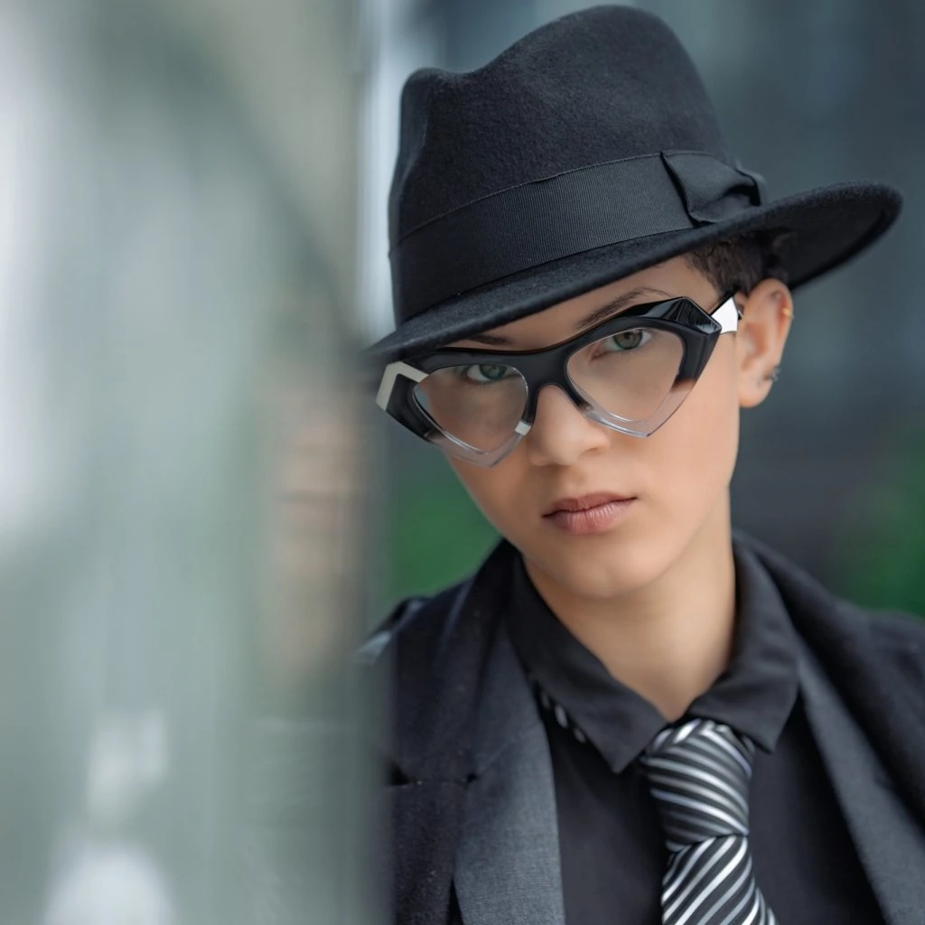 Riannah in a black fedora and angular black and clear Ffin NOS optical frames, striped tie, Tall Tales shoot