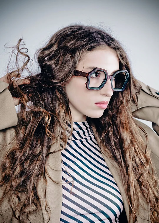 Woman with long wavy hair in hexagonal Ffin AER optical frames, hands in her hair, striped top