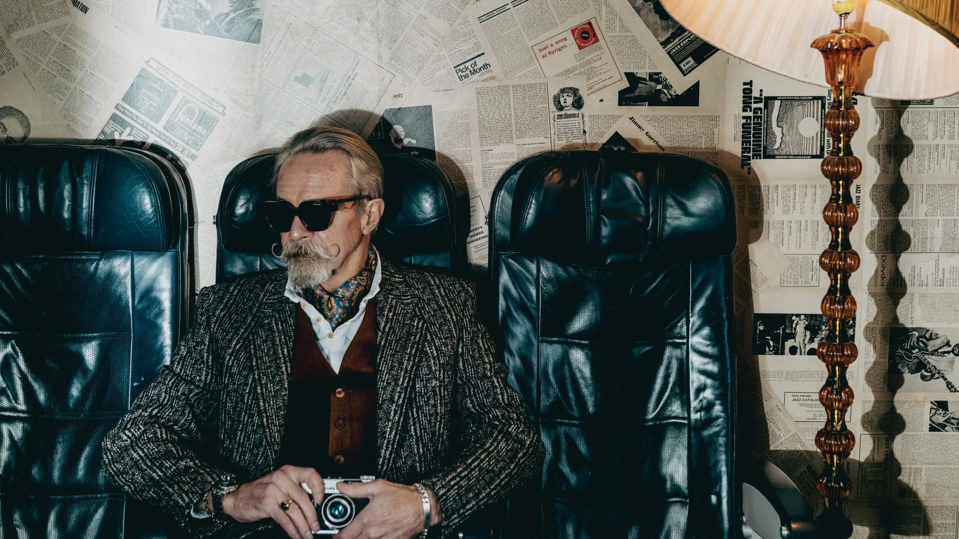 Gary in black Ffin AMO designer sunglasses seated between leather chairs holding a vintage camera
