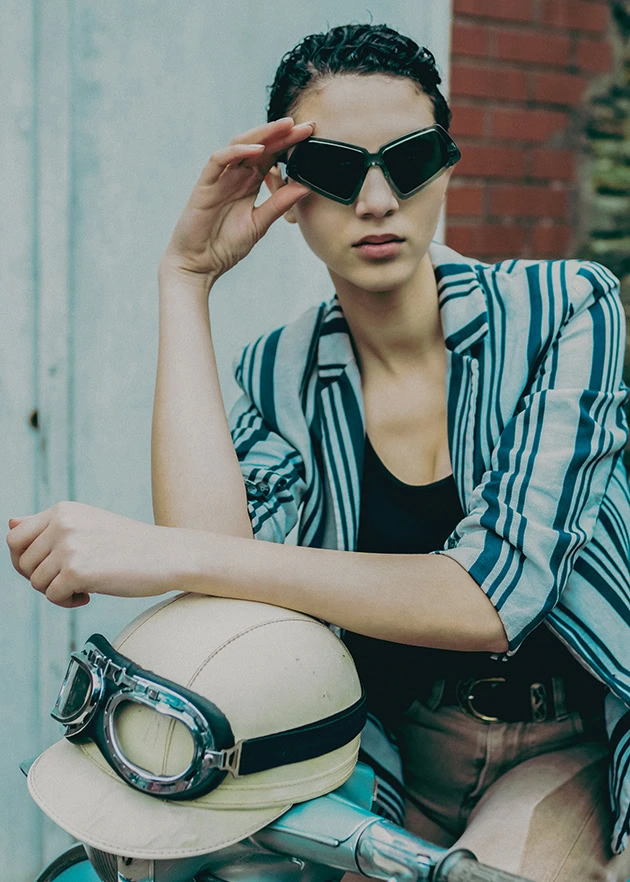 Model raising black angular Ffin MOD sunglasses in a striped blazer, beside a helmet and goggles