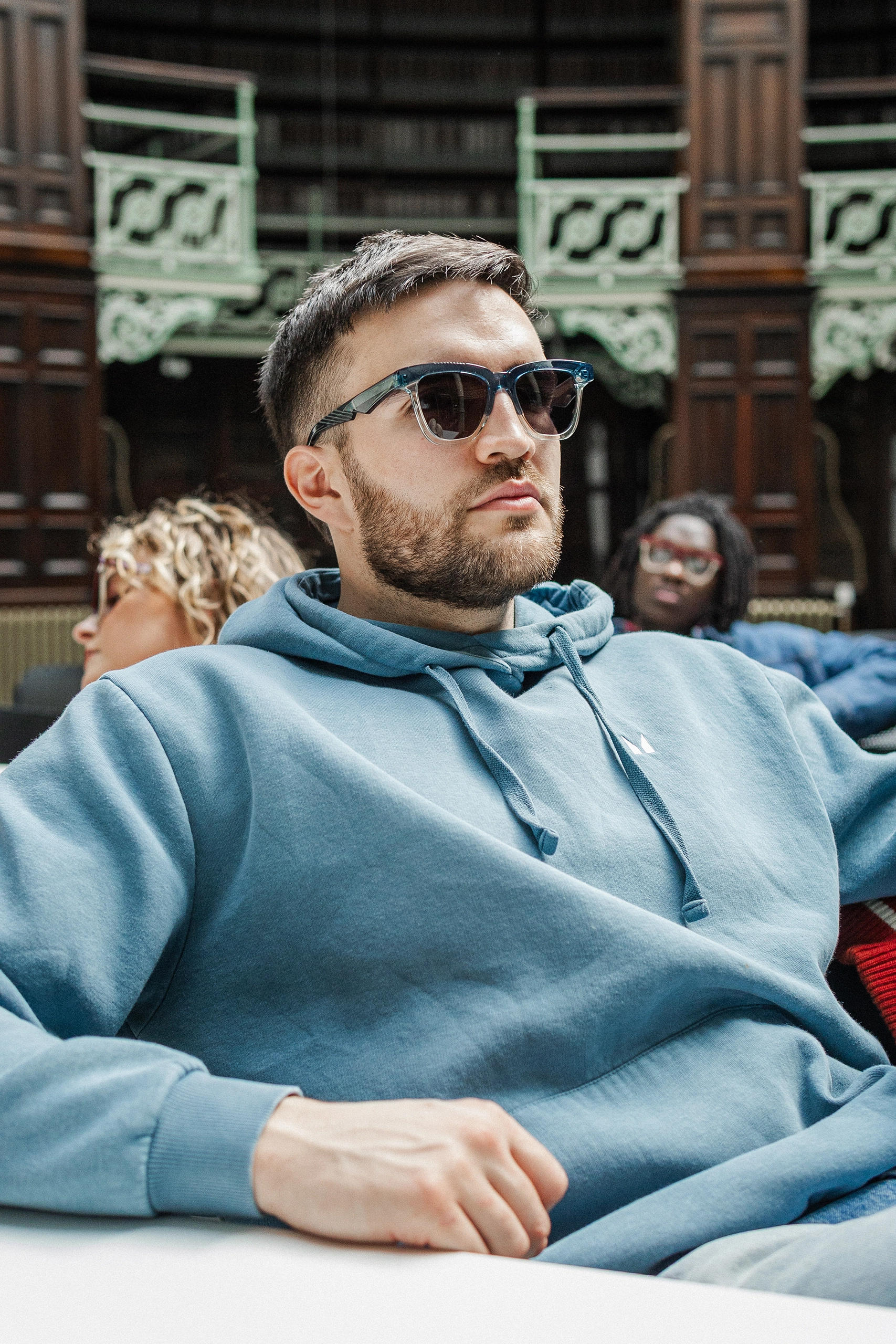 Man in a blue hoodie and Ffin GEN blue sunglasses in an ornate library, Breakfast Club shoot