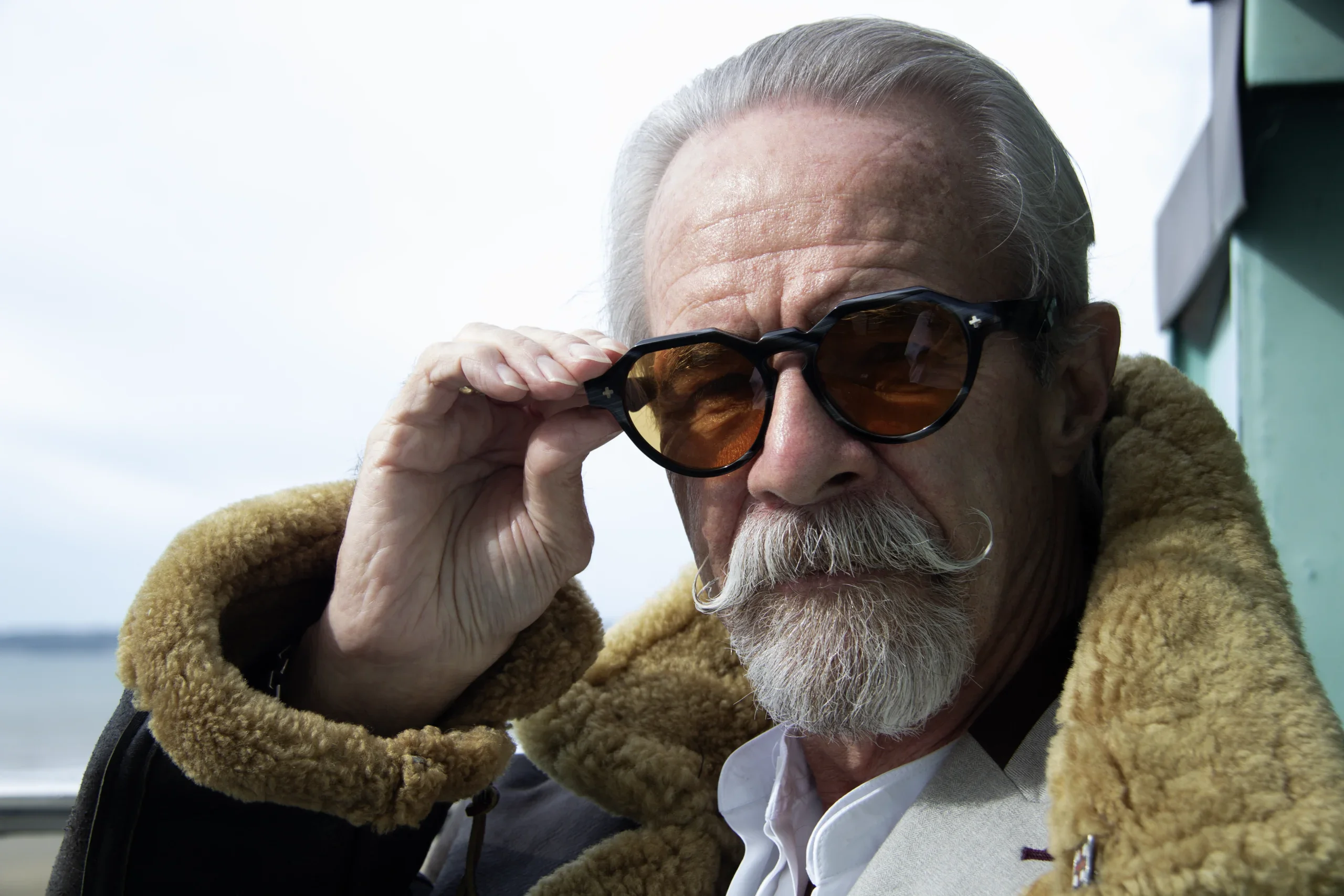 Gary lowering amber-lensed Ffin NON designer sunglasses, shearling collar, sea behind him