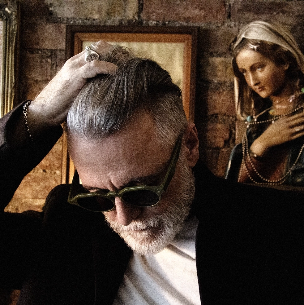 Steve Balsamo in green Ffin NON designer sunglasses running a hand through his hair beside a statue