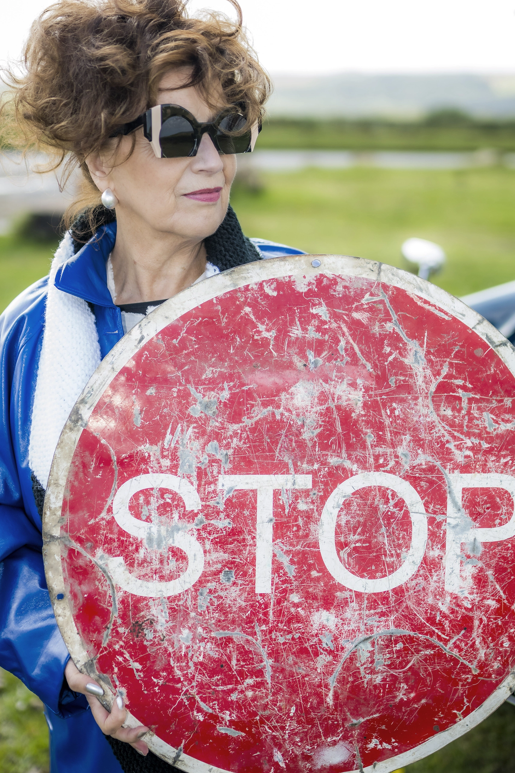 Rose in black angular Ffin ORA designer sunglasses holding a weathered STOP sign outdoors
