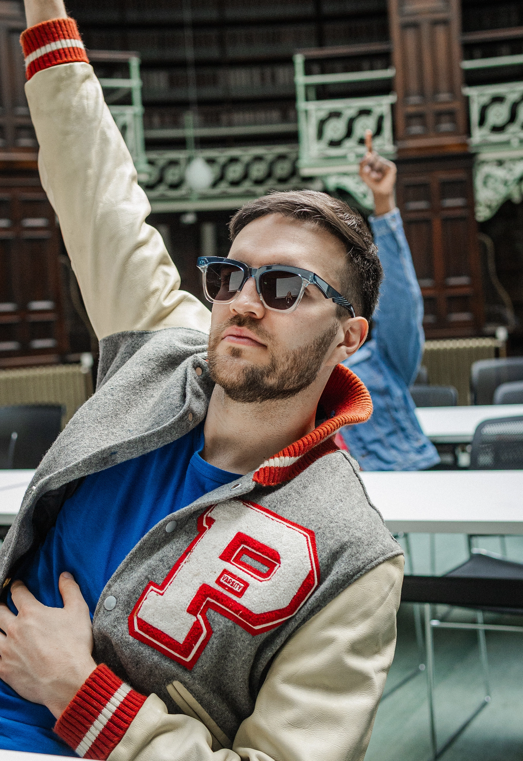 Man in a varsity jacket raising an arm in Ffin GEN blue sunglasses, library behind him
