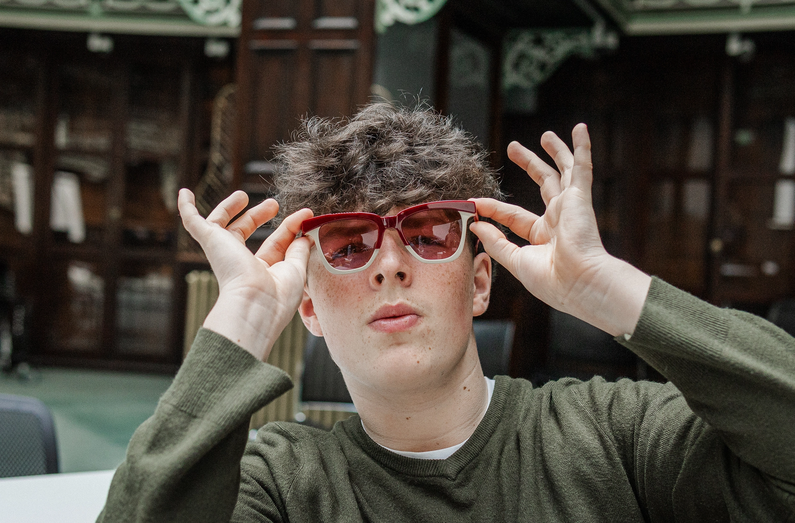 Young man adjusting red-tinted Ffin GEN designer sunglasses in a library