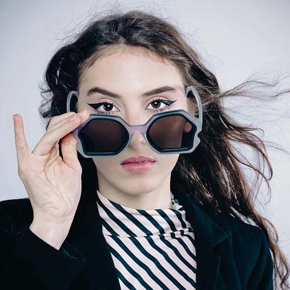 Woman lowering mauve hexagonal Ffin AER designer sunglasses, striped top and windswept hair